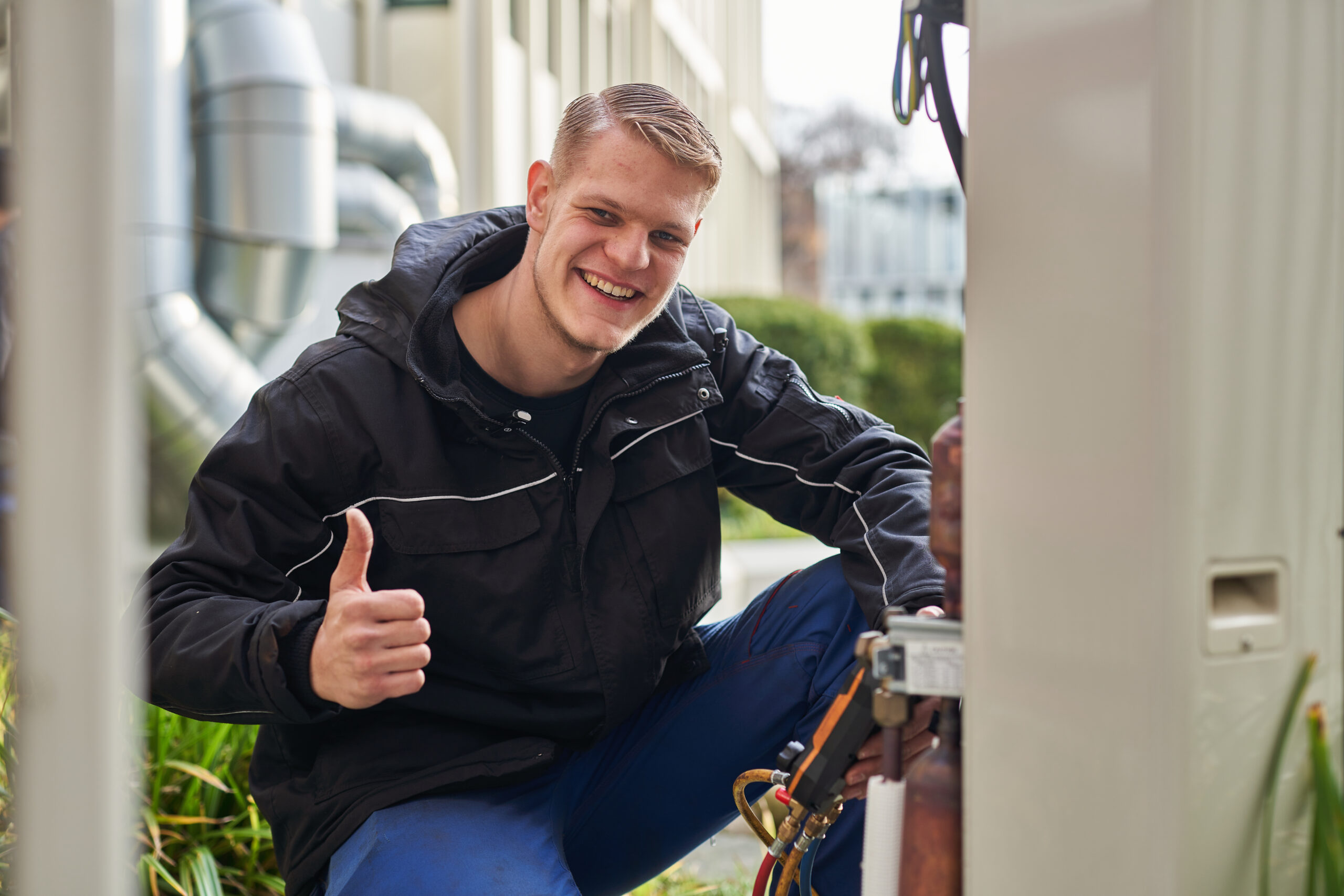 Ein junger Klimatechniker in dunkler Arbeitsjacke und blauer Arbeitshose hockt im Freien vor einer geöffneten Außeneinheit, blickt lächelnd in die Kamera und zeigt den Daumen nach oben. Mit der anderen Hand hält er ein Prüfwerkzeug mit Schläuchen, während im Hintergrund ein Industriegebäude mit dicken Lüftungsrohren zu sehen ist.