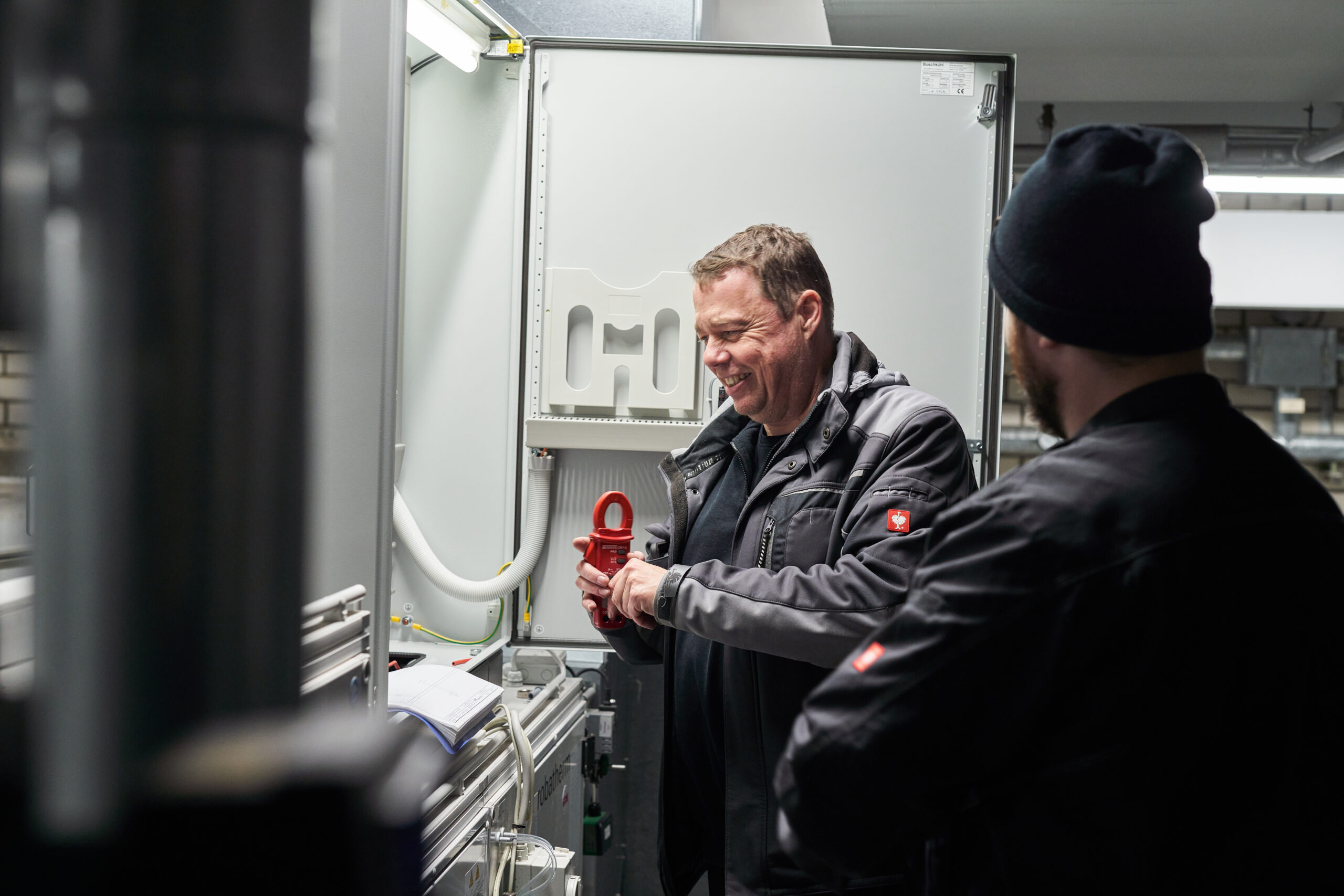 Zwei Techniker in grauer Arbeitskleidung arbeiten an einem geöffneten Schaltschrank in einem Technikraum. Der links stehende ältere Mann hält lächelnd eine rote Strommesszange in den Händen und unterhält sich gut gelaunt mit seinem Kollegen im Vordergrund, der mit schwarzer Mütze von hinten zu sehen ist.