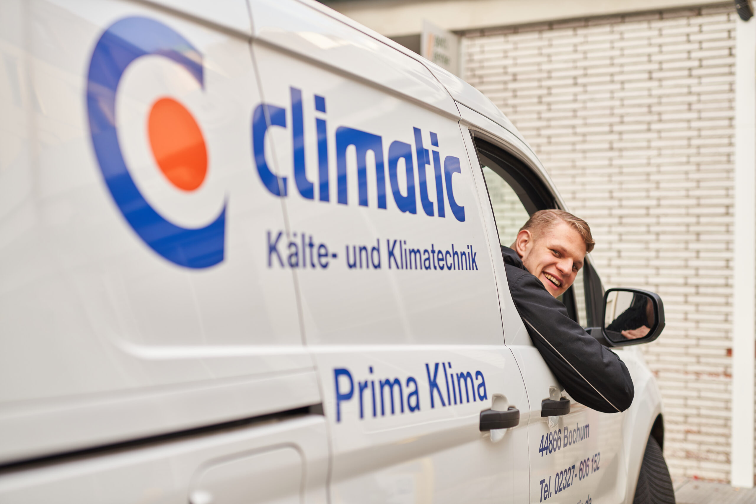 Ein lächelnder junger Mitarbeiter schaut aus dem geöffneten Seitenfenster eines weißen Firmenfahrzeugs des Unternehmens „climatic Kälte- und Klimatechnik“. Auf der Fahrzeugseite sind das Firmenlogo sowie der Schriftzug „Prima Klima“ und Kontaktdaten aus Bochum zu sehen.