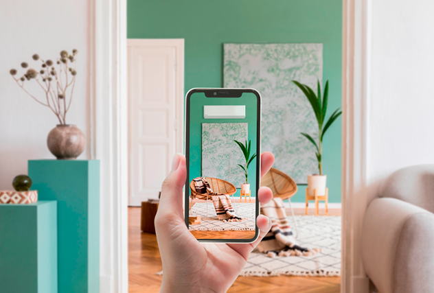 Eine Hand hält ein Smartphone hoch und nutzt eine Augmented-Reality-App zur Raumplanung, die ein weißes Klimagerät virtuell an die grüne Wand eines stilvollen Wohnzimmers projiziert. Der reale Raum im Hintergrund ist mit Holzfußboden, Sesseln, Teppich und Zimmerpflanzen eingerichtet.