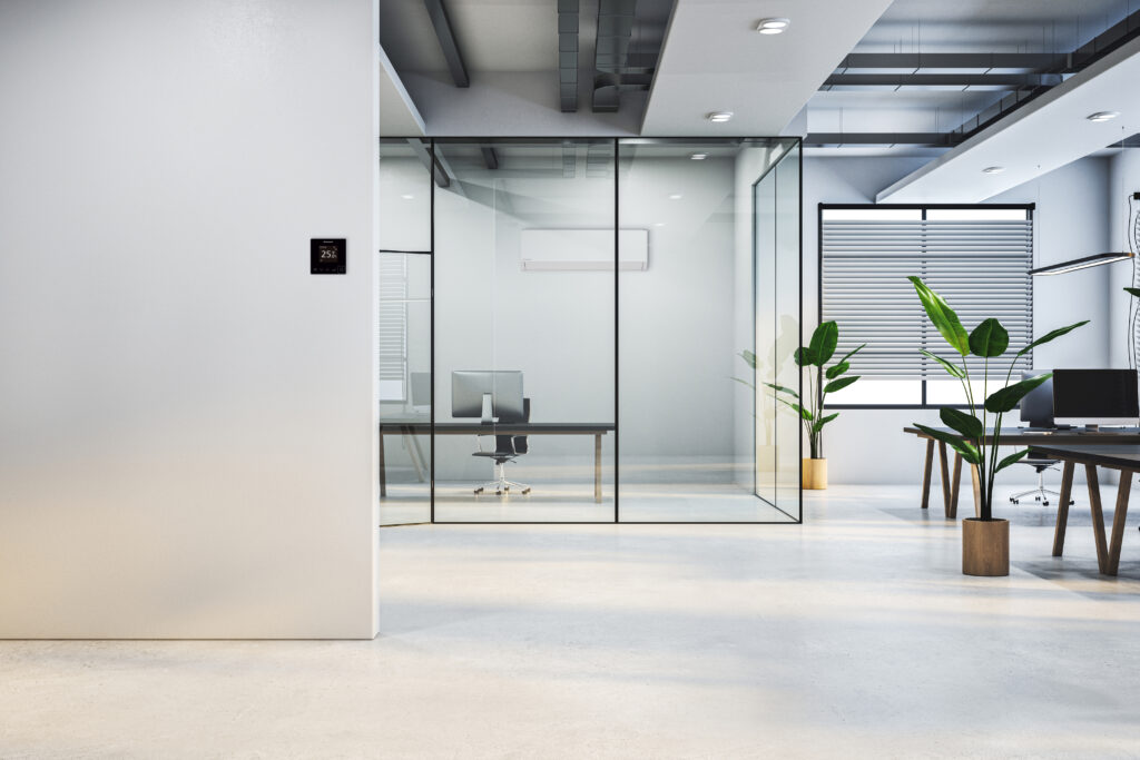 Alt-Text: Ein modernes, helles Großraumbüro im Loft-Stil mit hellem Betonboden, Glaskabinen und großen Topfpflanzen. An der weißen Wand im Vordergrund ist ein digitales Wandthermostat montiert, während man durch die Glasfronten in einen Besprechungsraum mit montiertem Klimagerät sowie rechts auf offene Arbeitsplätze mit Schreibtischen und Monitoren blickt.