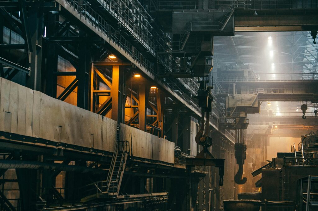 Das Innere einer riesigen, düsteren Industrie- oder Stahlwerkshalle mit massiven Stahlkonstruktionen, Laufstegen und Treppen. Von der Decke hängen schwere Kranhaken herab, während punktuelle gelbe Arbeitsleuchten und helles Licht im diesigen Hintergrund für eine stimmungsvolle Industrieatmosphäre sorgen.