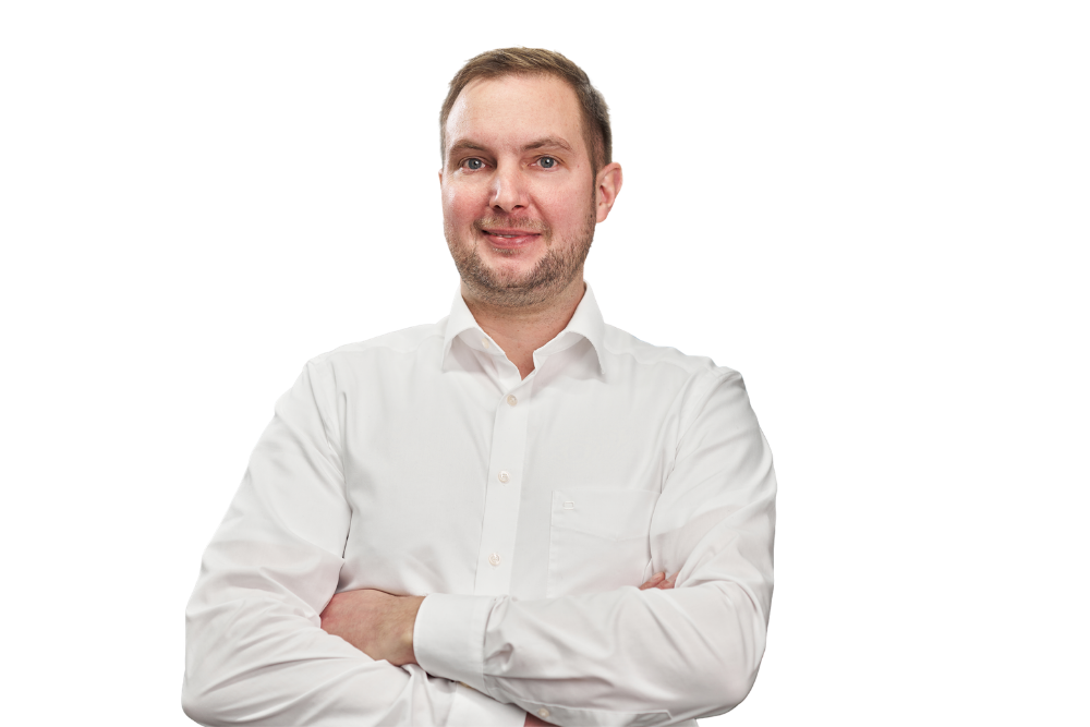 Ein lächelnder Mann mit kurzen braunen Haaren und leichtem Bart, der ein weißes Hemd trägt und die Arme vor der Brust verschränkt hat.