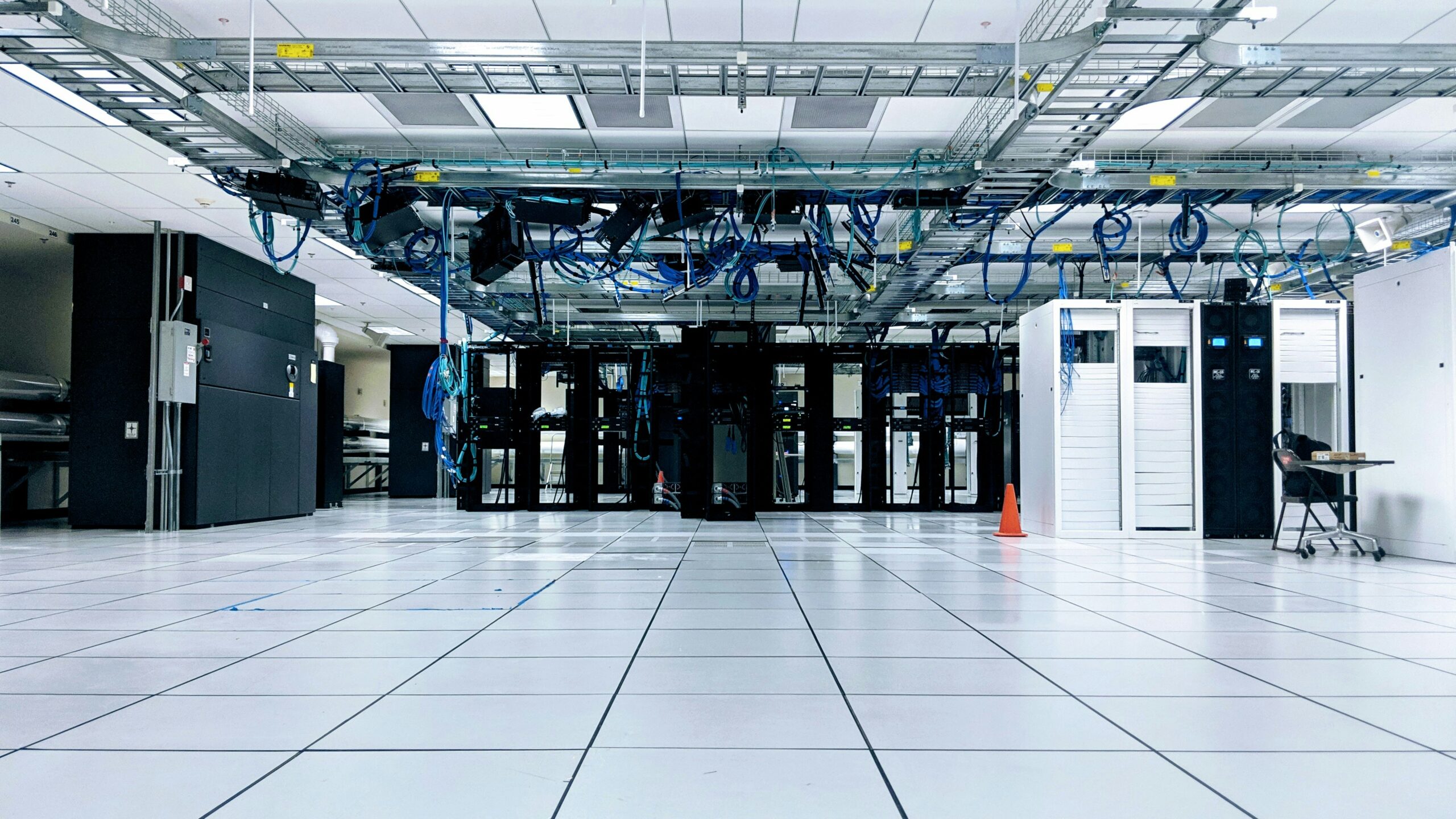 Ein geräumiges, modernes Rechenzentrum mit weißem Doppelboden und sichtbaren Kabeltrassen an der Decke, von denen dichte Bündel blauer Netzwerkkabel herabhängen. Im hinteren Bereich stehen Reihen schwarzer und weißer Serverschränke, davor steht ein kleiner orangefarbener Leitkegel auf dem Boden.